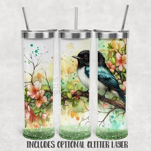Op de afbeelding: Drie ge&iuml;soleerde roestvrijstalen bekers met een vogel- en bloemenontwerp. De vogel heeft zwarte, witte en blauwgroene veren, gezeten op een tak met roze bloemen. De tekst onderaan luidt "INCLUDES OPTIONAL GLITTER LAYER."