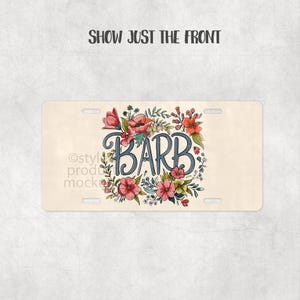 May include: A beige license plate featuring the name "BARB" in a blue, cursive font, encircled by a floral wreath of pink, red, and yellow flowers and green leaves. The text "SHOW JUST THE FRONT" is at the top.