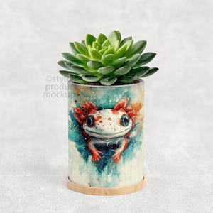 May include: A cylindrical planter pot featuring a watercolor axolotl design in shades of red, white, and blue. The pot holds a green succulent plant. The pot sits on a small, round, light brown base.