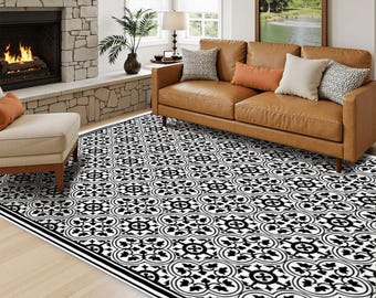 May include: A large rectangular area rug featuring a repeating black and white floral tile pattern with a solid black border. The rug is positioned on a light wood floor in a room with a stone fireplace, a tan sofa, and a matching ottoman.