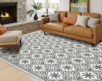 May include: A rectangular area rug featuring a repeating gray and white geometric pattern with stylized floral motifs. The rug covers a light-colored wooden floor in a living room setting, positioned in front of a brown leather sofa and a fireplace.