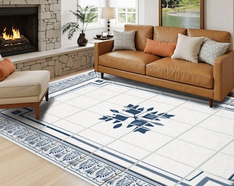 May include: A rectangular area rug featuring a white base with a grid pattern, a central blue geometric floral motif, and a decorative border with blue leaf and scroll designs. The rug is positioned on a light wood floor in a living room.