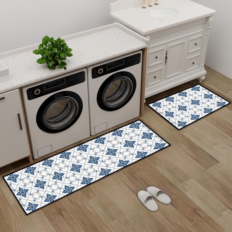 May include: Two patterned rugs with a blue floral design and black borders are placed on a wooden floor. The rugs are in front of a washing machine and a white cabinet. A small green plant sits on the countertop.