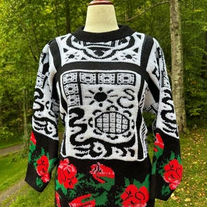 May include: A black, white, and red sweater with a crew neck. The sweater features a geometric pattern on the upper portion and red roses on the sleeves and lower hem. The sweater is displayed on a mannequin.