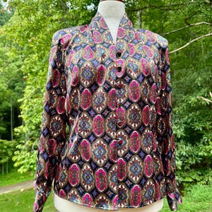 May include: A long-sleeved blouse with a vibrant, repeating pattern of pink, blue, and brown shapes on a dark blue background. The blouse has a mandarin collar and is buttoned down the front. The garment is displayed on a mannequin against a backdrop of green foliage.