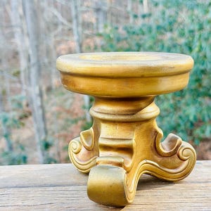 Op de afbeelding: Een goudkleurige, sierlijke sokkel met een ronde bovenkant en decoratief krulwerk. De sokkel heeft een stevige basis en wordt waarschijnlijk gebruikt voor het tentoonstellen van decoratieve items. De achtergrond toont een wazig zicht op bomen.