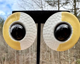 May include: A pair of round, white and yellow ceramic earrings with a black center. The earrings have a textured surface and a raised design.