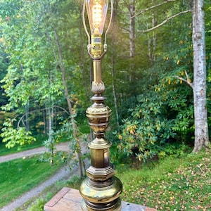 Puede incluir: Una lámpara de mesa de latón vintage con una bombilla estilo Edison. La lámpara tiene un diseño detallado y ornamentado con una base redonda y un cuerpo alto y delgado. La bombilla está encendida, emitiendo un brillo cálido. La lámpara está sobre una superficie de madera.