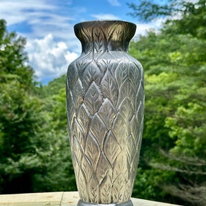 Könnte beinhalten: Eine silberfarbene Metallvase mit einem durchgehenden, geprägten Blattmuster auf der gesamten Außenfläche. Die Vase hat einen schmalen Hals, der sich am oberen Rand leicht weitet, sowie einen runden Fuß und eine polierte Oberfläche.