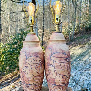May include: Two ceramic table lamps featuring a Native American profile design. The lamps are brown and tan, with gold-coloured lamp holders and vintage-style light bulbs. The lamps are approximately 51 cm tall.