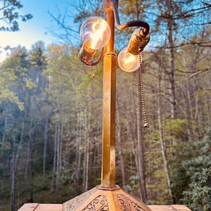 May include: An antique brass lamp with two illuminated Edison-style bulbs. The lamp has a decorative base with an ornate design and a central column. The background features a blurred view of trees and a blue sky.