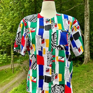 Könnte beinhalten: Ein kurzärmeliges Hemd mit einem lebendigen, geometrischen Muster. Das Design zeigt vertikale Streifen in Grün, Weiß und Blau, durchsetzt mit roten, gelben und schwarzen Formen. Das Hemd wird auf einer Schaufensterpuppe in einer natürlichen Umgebung präsentiert.