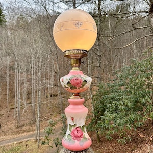 May include: An antique table lamp with a pink and white painted design featuring roses. The lamp has a round, frosted glass globe and a brass base. The lamp stands approximately 30 inches tall.