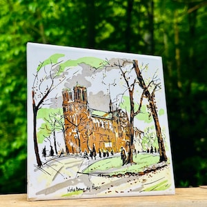 May include: A square ceramic tile featuring a detailed ink and watercolor illustration of the Notre Dame de Paris cathedral. The artwork depicts the building in shades of brown, accented by green foliage and black outlines, with the text Notre Dame de Paris written in black ink at the bottom.