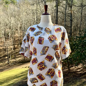 Könnte beinhalten: Weißes Kurzarm-Oberteil mit einem sich wiederholenden Muster aus offenen Büchern. Die Bücher haben rote, gelbe und violette Illustrationen. Die Bluse wird auf einer Schaufensterpuppe vor einem natürlichen Hintergrund im Freien präsentiert.