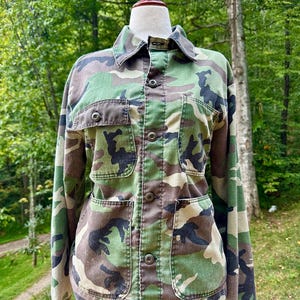 May include: Camouflage pattern jacket with button-up front, two chest pockets, and two lower pockets. The jacket is displayed on a mannequin. The colors include shades of green, brown, and black. The jacket has long sleeves and a collared neckline.