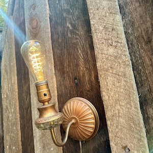 May include: Vintage-style wall sconce with a decorative brass finish. The sconce features a clear glass shade and a curved arm, holding a vintage-style light bulb. The fixture is mounted on a wooden surface, creating a rustic aesthetic.