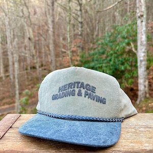 Op de afbeelding: Een pet met een blauwe klep en een lichtgroene bovenkant. De tekst "HERITAGE GRADING & PAVING" is op de voorkant geborduurd. Een gevlochten touwdetail scheidt de klep van de bovenkant.