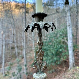 Puede incluir: Un candelabro antiguo de latón con una vela de color crema. El candelabro presenta diseños ornamentados de hojas y flores. El fondo es una vista borrosa de un bosque con árboles desnudos.
