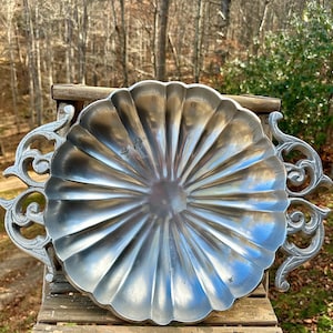 May include: A large, silver-colored metal serving tray with a scalloped edge and ornate, scroll-like handles. The tray is placed on a wooden surface, with a natural outdoor background of trees and foliage.
