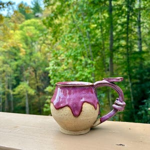 May include: A ceramic mug with a rounded base and a glossy magenta glaze dripping over the upper portion, while the lower section remains a natural beige clay. The handle features a decorative knot detail and a flat thumb rest at the top.