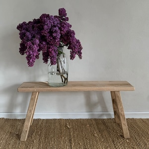 May include: A wooden bench with angled legs holds a clear glass vase overflowing with purple lilacs. The bench is made of light-coloured wood and sits on a woven jute rug. The bench is approximately 100 cm long.