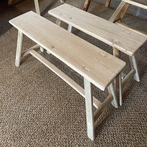 Puede incluir: Dos bancos de madera clara con asientos rectangulares y patas anguladas. Los bancos están hechos de madera sin terminar, mostrando la veta natural. Cada banco tiene tres hendiduras ovaladas en el asiento. Los bancos están colocados sobre una alfombra tejida y texturizada.