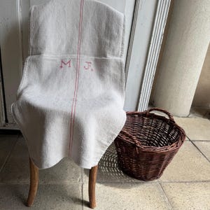 May include: A beige linen towel featuring a vertical red stripe and the embroidered red initials M J, draped over a wooden chair next to a round, dark brown woven wicker basket with two handles. The fabric has a visible, coarse-woven texture.