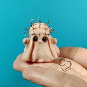 May include: A small, white, handmade doll with a stitched-up face and a crown of silver pins. The doll is wearing a white, flowing gown and is being held in a hand.