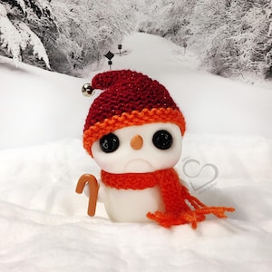 May include: A snowman figure wearing a red and orange knitted hat and scarf, and holding a candy cane. The snowman has large black eyes and a sad expression. The background is a snowy winter scene.