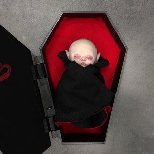 Pode incluir: Um pequeno boneco de bebê pálido com os olhos fechados e uma pequena boca está deitado em um caixão de madeira preto com um forro de veludo vermelho. O boneco está usando uma capa preta.