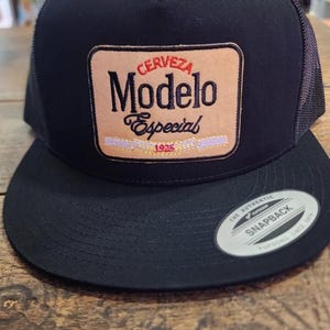 May include: Black snapback hat with a mesh back and a flat brim. The front features a tan patch with the words "CERVEZA Modelo Especial 1925" in red and black lettering. A white sticker is on the brim.