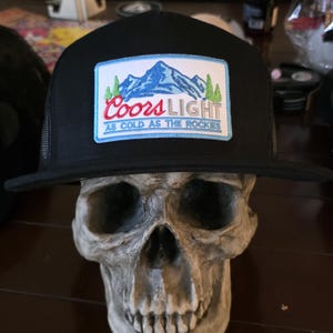 May include: Black trucker hat with a rectangular patch featuring a mountain scene and the words "Coors Light" in red and white. The text "As Cold As The Rockies" is below the logo. The hat is displayed on a skull.