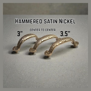 Könnte beinhalten: Drei Schrankgriffe aus gehämmertem Satin-Nickel. Die Griffe sind gewölbt mit einer strukturierten Oberfläche und dekorativen Enden. Der Text oben lautet "Hammered Satin Nickel" und "Center to Center". Die Griffe sind 7,6 cm und 8,9 cm von Mitte zu Mitte.