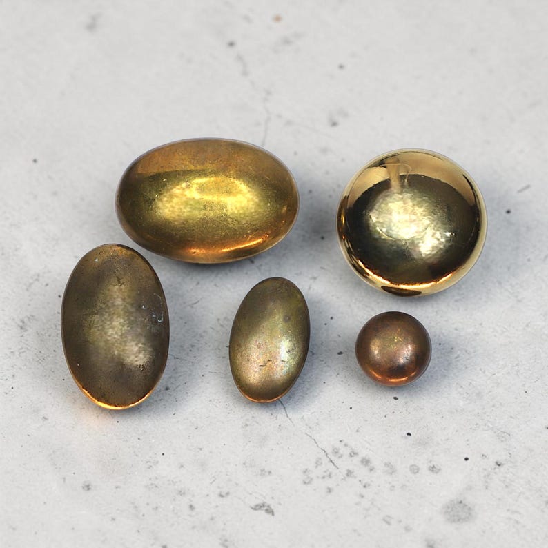May include: A collection of six metallic buttons in different shapes and sizes. The buttons vary in colour from gold to bronze, with oval and round forms. The buttons are displayed on a light grey surface.