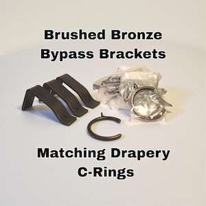 Puede incluir: Soportes de derivación de bronce cepillado y anillas en C a juego para cortinas. La imagen muestra varios soportes, una anilla en C y un paquete de anillas. El texto de la imagen dice "Brushed Bronze Bypass Brackets" y "Matching Drapery C-Rings."
