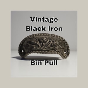 May include: Vintage black iron bin pull with a decorative floral design. The bin pull is a dark, matte black color and has two holes for mounting. The text "Vintage Black Iron Bin Pull" is displayed above and below the pull.