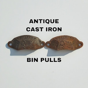 Op de afbeelding: Twee antieke gietijzeren bakgrepen met een verweerde, roestige afwerking. De grepen hebben een decoratief, geometrisch ontwerp. De tekst luidt "ANTIQUE CAST IRON BIN PULLS".