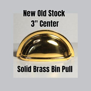 May include: A solid brass bin pull with a 3-inch center, new old stock. The pull has a curved, half-moon shape and a shiny gold finish. The text "New Old Stock 3" Center" and "Solid Brass Bin Pull" are displayed in black.