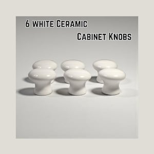 May include: Six white ceramic cabinet knobs are arranged in a row. The knobs are a classic mushroom shape with a glossy finish. The text "6 WHITE CERAMIC CABINET KNOBS" is displayed above the knobs.