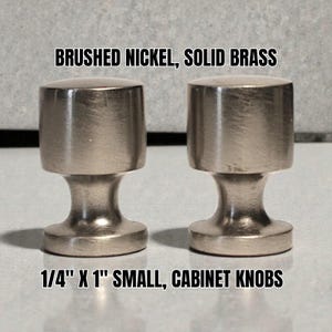 Pode incluir: Dois puxadores de armário em níquel escovado. Cada puxador tem cerca de 2,5 cm de comprimento e 0,6 cm de largura. Os puxadores são feitos de latão maciço. O texto na imagem diz: "BRUSHED NICKEL, SOLID BRASS" e "1/4" X 1" SMALL, CABINET KNOBS."