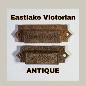 以下が含まれることがあります： 2つのアンティークイーストレイクビクトリア朝の引き出しプル。プルは長方形で、華やかな花柄と幾何学模様が施されています。「Eastlake Victorian」という言葉が上にあり、「ANTIQUE」という言葉が下にあります。