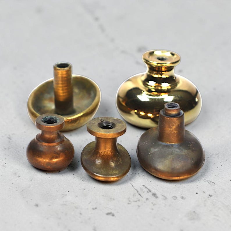 May include: Five antique cabinet knobs in various shapes and finishes. The knobs are made of brass and bronze, with a mix of polished gold and aged patina. The shapes include a mushroom, a rounded oval, and a cylindrical design.