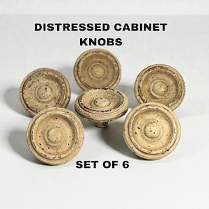 May include: A set of six distressed cabinet knobs. The knobs are round with a raised center and a textured, aged appearance. The color is a mix of beige and brown, with darker speckles. The text "DISTRESSED CABINET KNOBS" and "SET OF 6" are visible.
