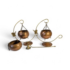 May include: A set of three copper-colored metal hanging bowls with decorative brass-colored supports. The bowls are suspended by chains from curved arms, with small angel figures adorning the supports. The design includes a chain and decorative elements.