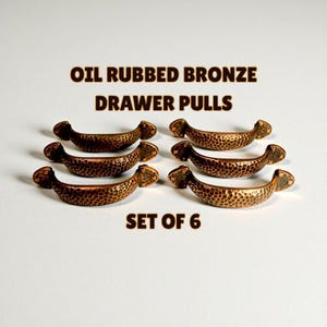 Puede incluir: Juego de seis tiradores de cajón de bronce aceitado. Cada tirador tiene una textura martillada y un diseño curvo. El texto "OIL RUBBED BRONZE DRAWER PULLS" y "SET OF 6" se muestra en la imagen.