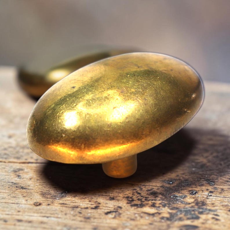 May include: Close-up of two antique brass cabinet knobs with an oval shape. The knobs have a warm, golden hue and a slightly aged appearance, suggesting they are vintage or vintage-inspired. They are set against a rustic wooden surface.