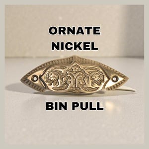 Puede incluir: Tirador de cubo de níquel adornado con un intrincado diseño floral. El herraje tiene un patrón decorativo y simétrico con dos orificios para el montaje. El texto "ORNATE NICKEL BIN PULL" se muestra en negrita negra.
