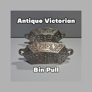 May include: Two antique Victorian bin pulls. The pulls are silver-colored, with intricate geometric and floral designs. The text "Antique Victorian" and "Bin Pull" are displayed above and below the pulls.