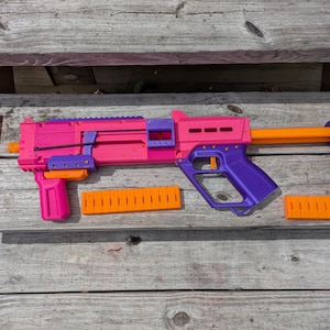 May include: A brightly colored toy gun in shades of pink, purple, and orange, with additional orange components. The toy gun is lying on a weathered wooden surface, with several orange accessories nearby.